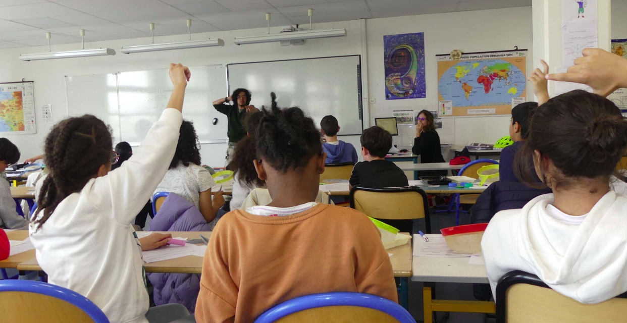 DC Anass Butsch is standing in the far background while teaching a class of kids seen in the foreground with their backs to the camera. 