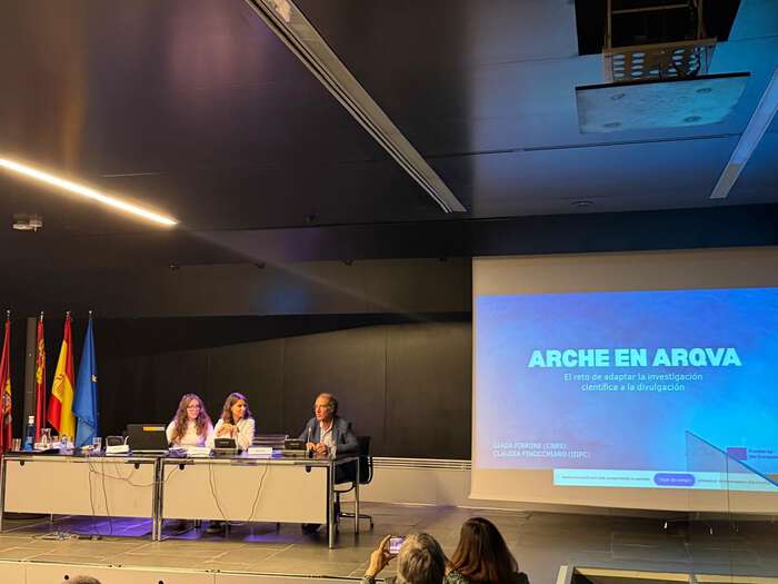 Giada Pirrone, Claudia Finocchiaro and Pablo Arias are sitting at a podium on a stage. To the right is a screen displaying a presentation.