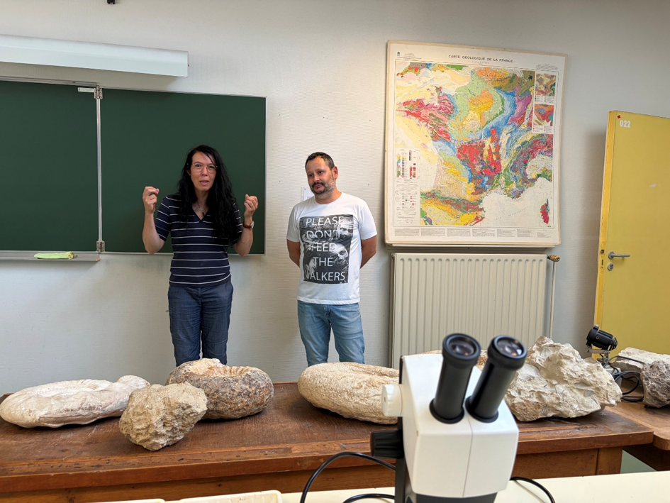 Two people, Catherine Dupont and Esteban Álvarez-Fernández, are standing in front of a white board. In front of them are several large rocks placed on a wooden table. It the very foreground in the centre is a microscope.